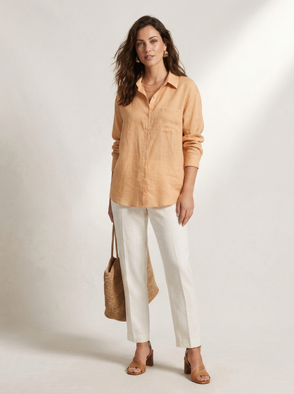 A woman stands against a light neutral studio background wearing a light peach button-up shirt, white straight-leg pants, tan heeled sandals, and holding a woven straw tote bag.