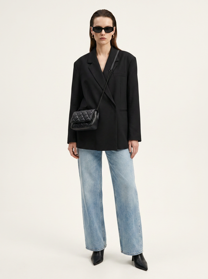 A model stands against a plain light background wearing a black double-breasted blazer, wide-leg light-wash blue jeans, black pointed shoes, and a black quilted shoulder bag.
