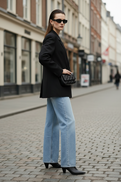 A woman stands on a cobblestone street wearing a black oversized blazer, light-wash wide-leg jeans, black ankle boots, and black sunglasses, with a black quilted shoulder bag.