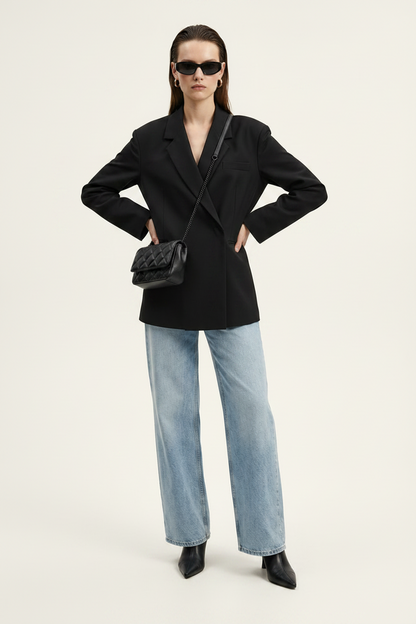 A model stands against a plain light background wearing a black double-breasted blazer, wide-leg light-wash blue jeans, black pointed shoes, and a black quilted shoulder bag.