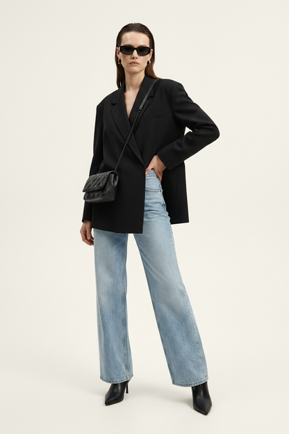A model stands against a plain light background wearing a black double-breasted blazer, wide-leg light-wash blue jeans, black pointed shoes, and a black quilted shoulder bag.
