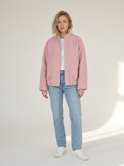 A woman modeling a light pink textured jacket with a stand collar, worn over a white t-shirt and light blue jeans, standing against a warm off-white studio background.