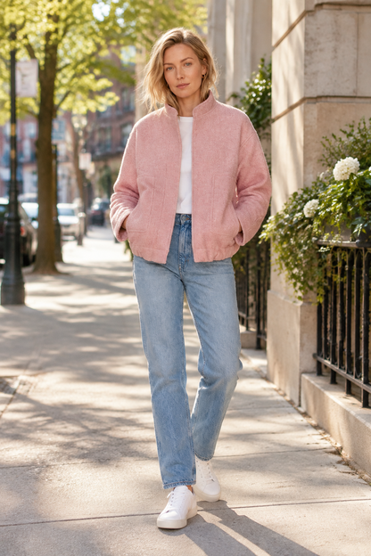 A woman stands on a sunny city sidewalk wearing a light pink zip-front jacket over a white top, light-wash blue jeans, and white sneakers.