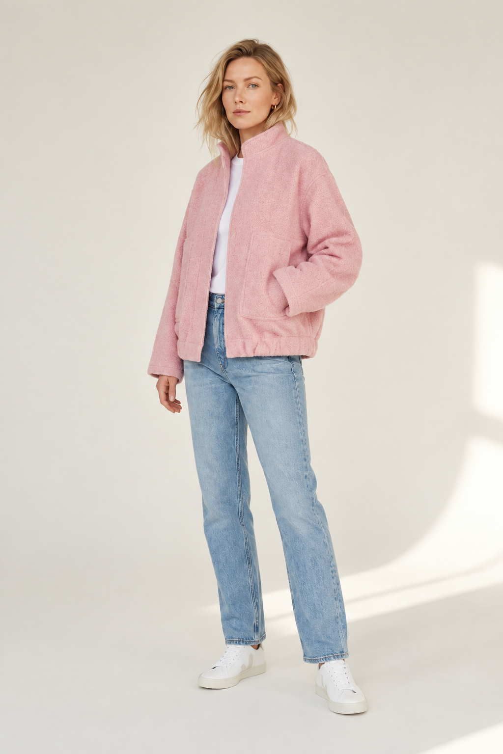 A woman modeling a light pink textured zip-up jacket, paired with light-wash blue jeans, a white top, and white sneakers, standing against a neutral off-white studio background.