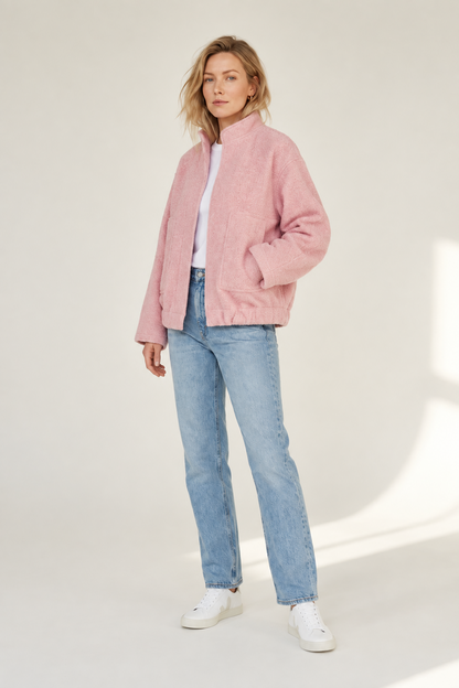 A woman modeling a light pink textured zip-up jacket, paired with light-wash blue jeans, a white top, and white sneakers, standing against a neutral off-white studio background.