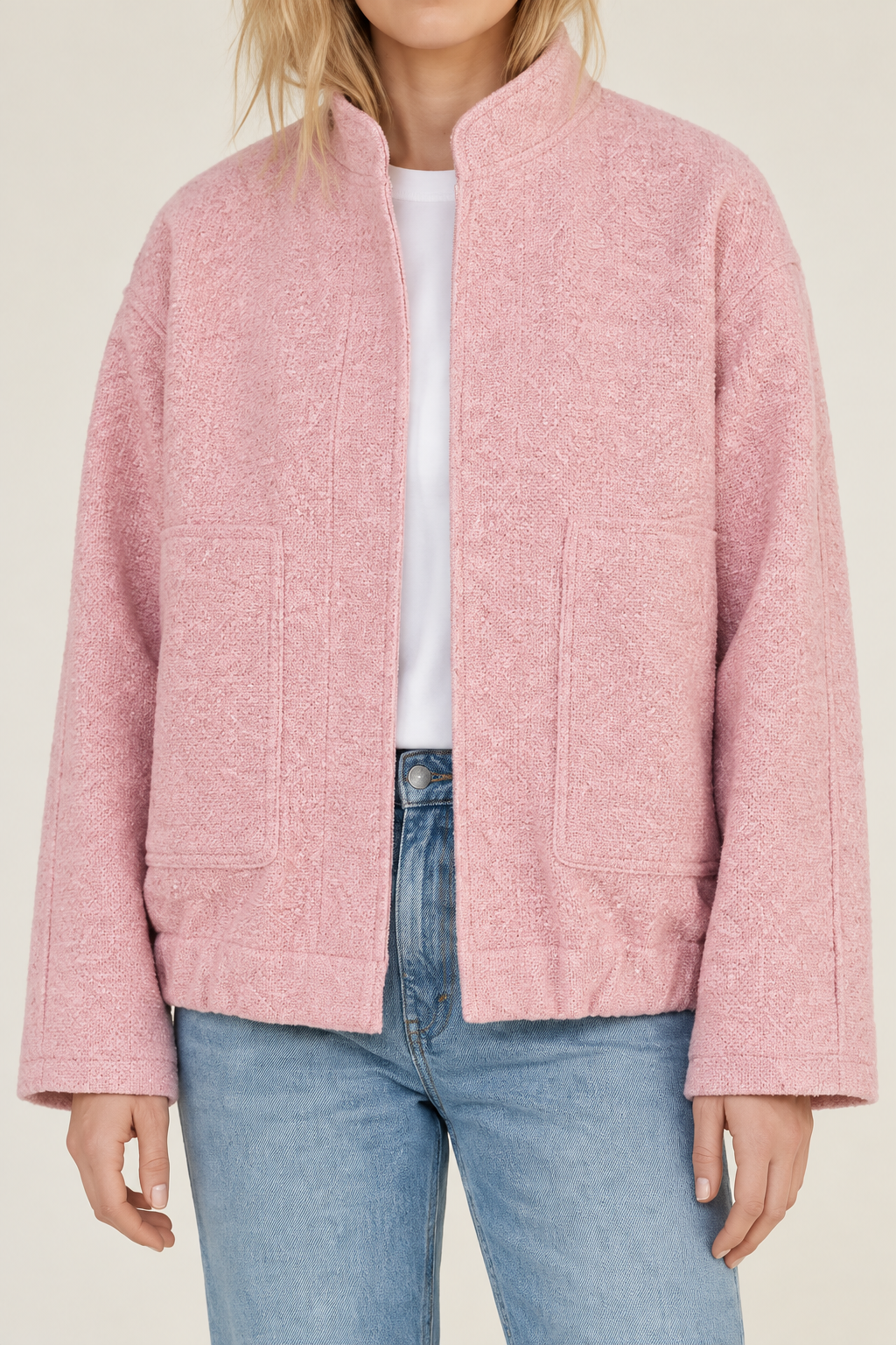 A model wearing a light pink textured bouclé jacket with a stand collar, front zipper, and two front patch pockets, styled over a white t-shirt and light blue jeans.