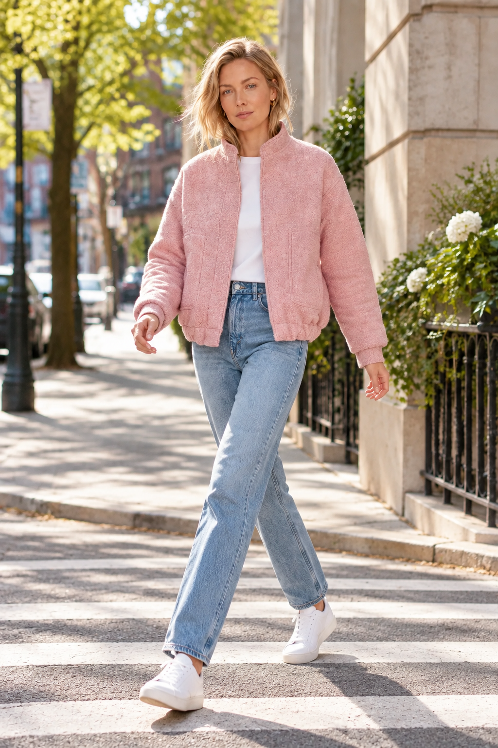 A woman walking on a city crosswalk wearing a fuzzy light pink bomber jacket, light blue jeans, and white sneakers.