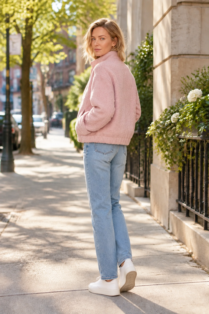 A woman stands on a city sidewalk wearing a light pink fuzzy cropped jacket, light blue jeans, and white sneakers, photographed in a sunny urban setting with trees and a stone building in the background.
