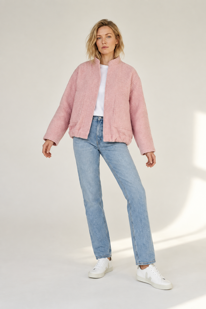 A woman modeling a light pink textured jacket, light-wash blue jeans, a white t-shirt, and white sneakers against a neutral studio backdrop.