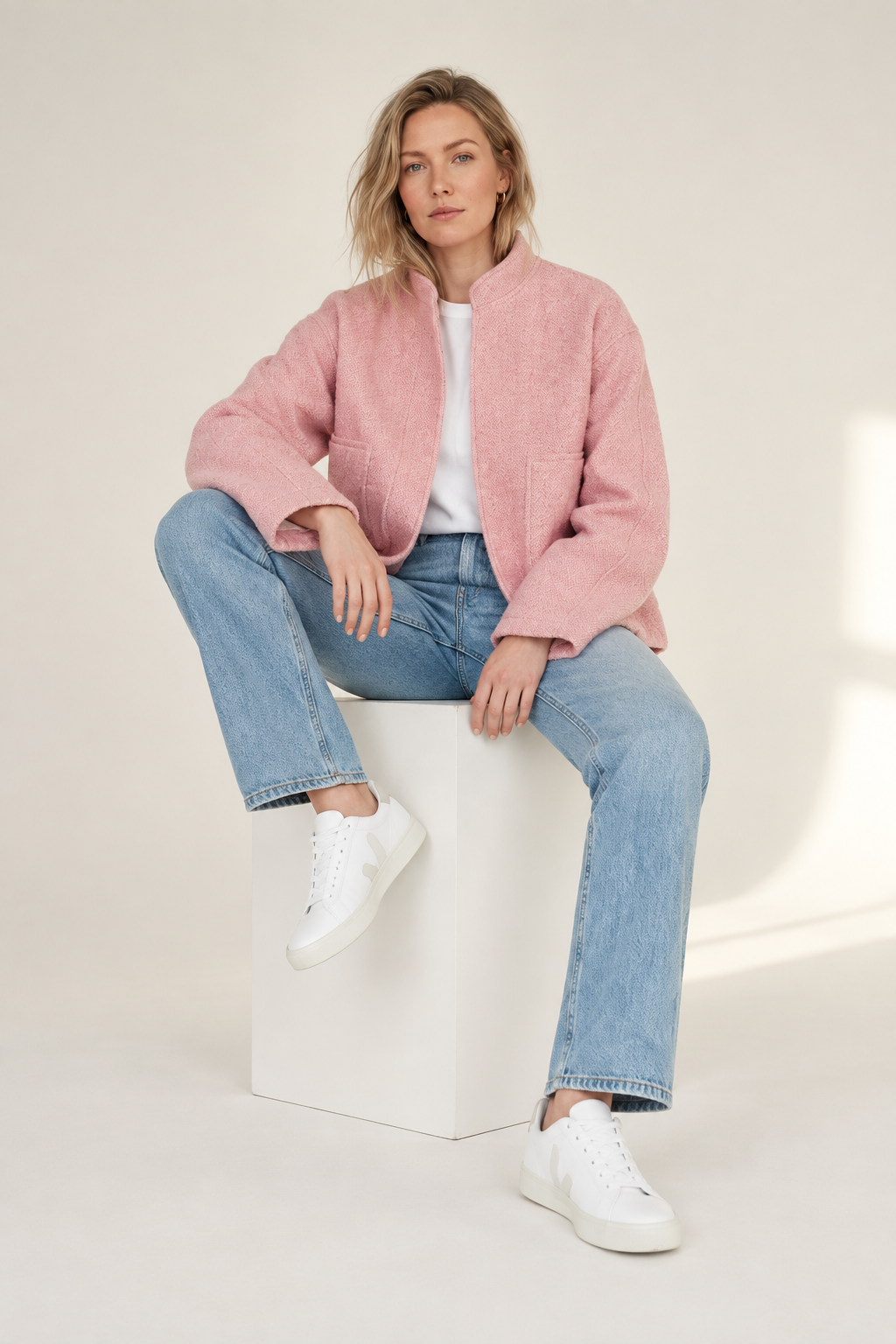 A woman sits on a white cube in a studio setting, wearing a pink fuzzy cropped jacket over a white top, light blue jeans, and white sneakers.