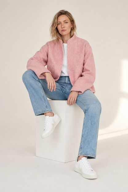 A woman sits on a white cube in a studio setting, wearing a pink fuzzy cropped jacket over a white top, light blue jeans, and white sneakers.