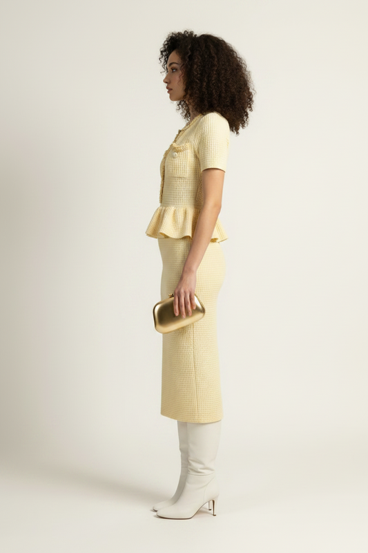 A model stands against a plain light background wearing a pale yellow short-sleeve knit top and matching textured midi skirt, paired with white knee-high boots and holding a small metallic clutch.
