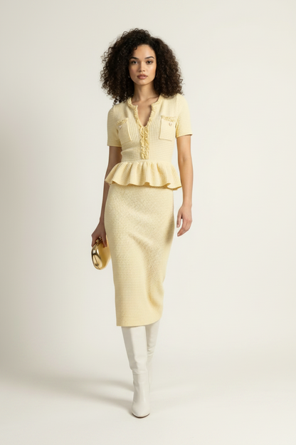 A model stands against a plain light background wearing a pale yellow short-sleeve knit top and matching textured midi skirt, paired with white knee-high boots and holding a small metallic clutch.