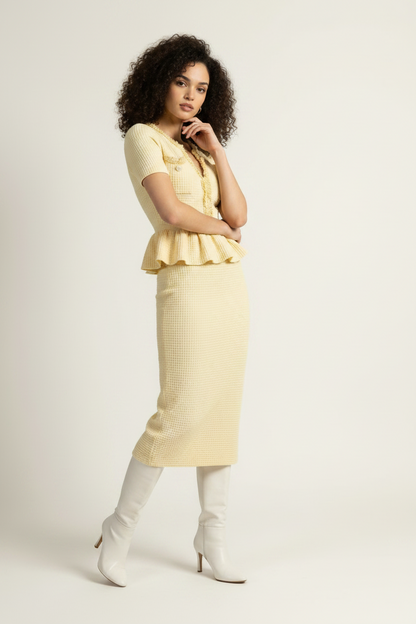 A model stands against a plain light background wearing a pale yellow short-sleeve knit top and matching textured midi skirt, paired with white knee-high boots and holding a small metallic clutch.