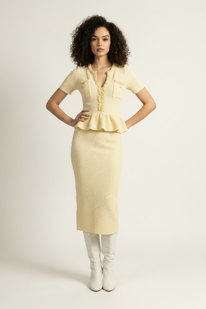 A model stands against a plain light background wearing a pale yellow short-sleeve knit top and matching textured midi skirt, paired with white knee-high boots and holding a small metallic clutch.