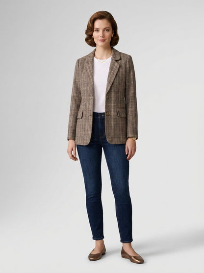A woman stands against a light gray studio background wearing a brown and beige plaid blazer over a white top, dark blue skinny jeans, and brown flat shoes.