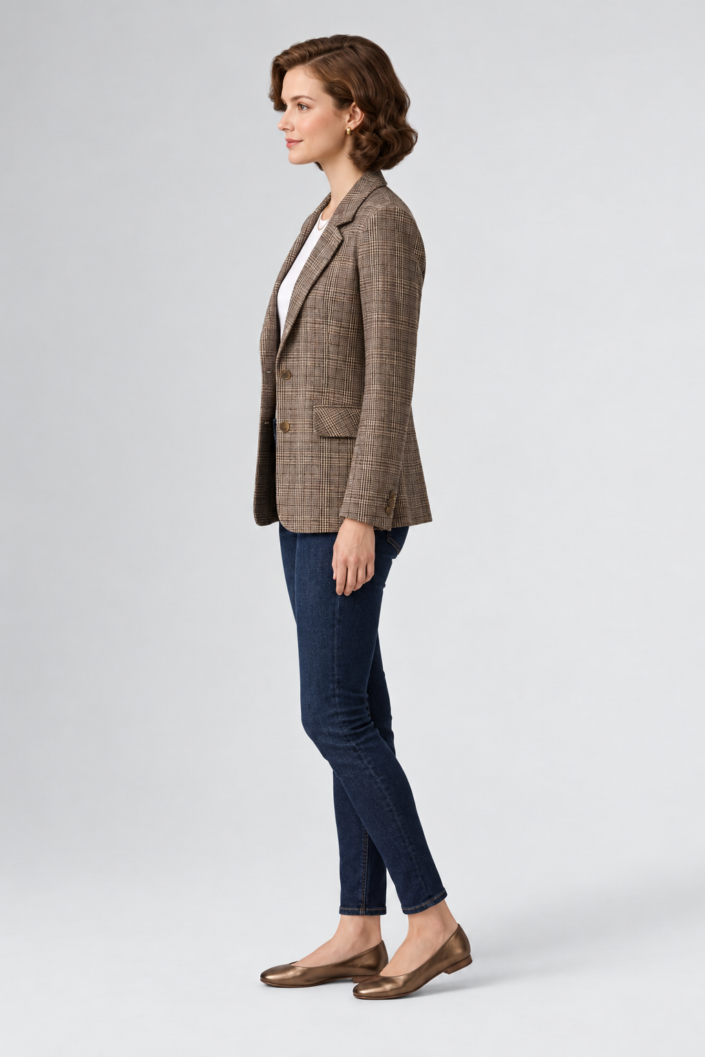 A woman stands against a light gray studio background wearing a brown and beige plaid blazer over a white top, dark blue skinny jeans, and brown flat shoes.