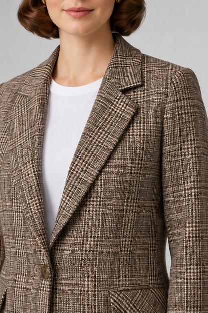 A woman stands against a light gray studio background wearing a brown and beige plaid blazer over a white top, dark blue skinny jeans, and brown flat shoes.