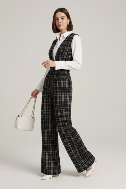 A model stands against a light gray studio background wearing a black and white plaid wide-leg vest and matching trousers over a white button-up shirt, holding a small white handbag.
