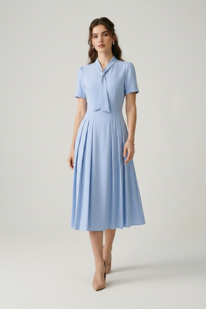 A woman modeling a light blue midi dress with short sleeves, a tie-neck detail, and a pleated skirt, paired with beige heels, against a neutral studio background.