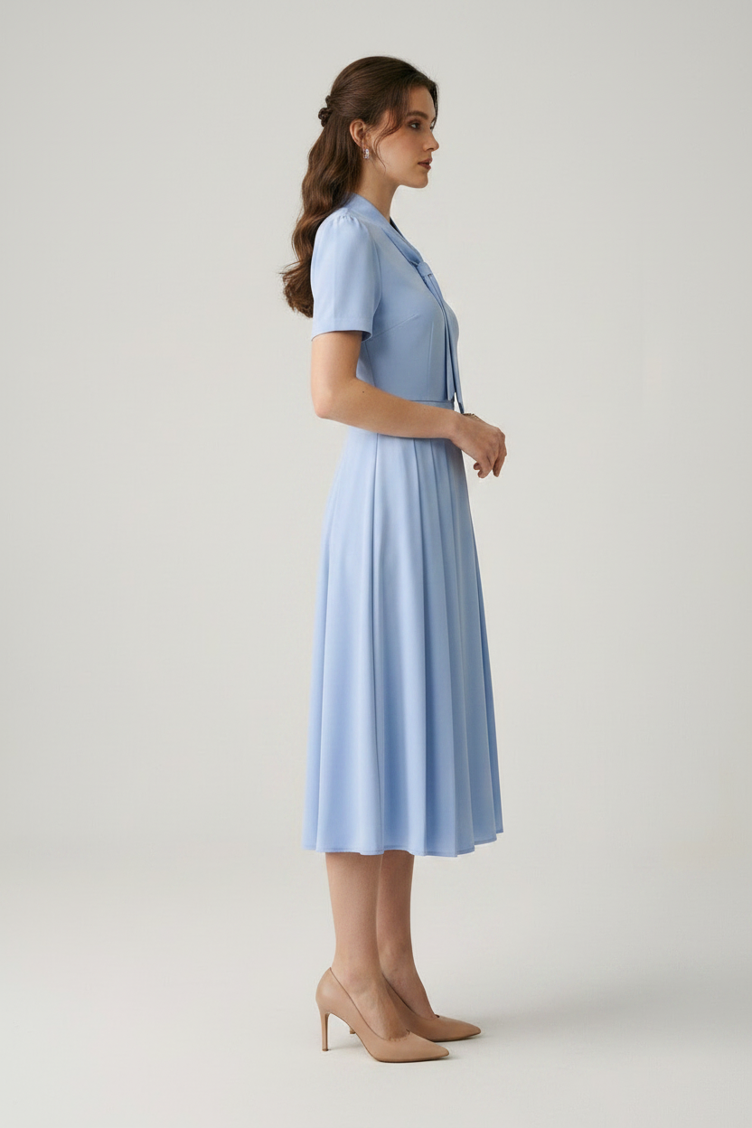 A woman modeling a light blue midi dress with short sleeves, a tie-neck detail, and a pleated skirt, paired with beige heels, against a neutral studio background.