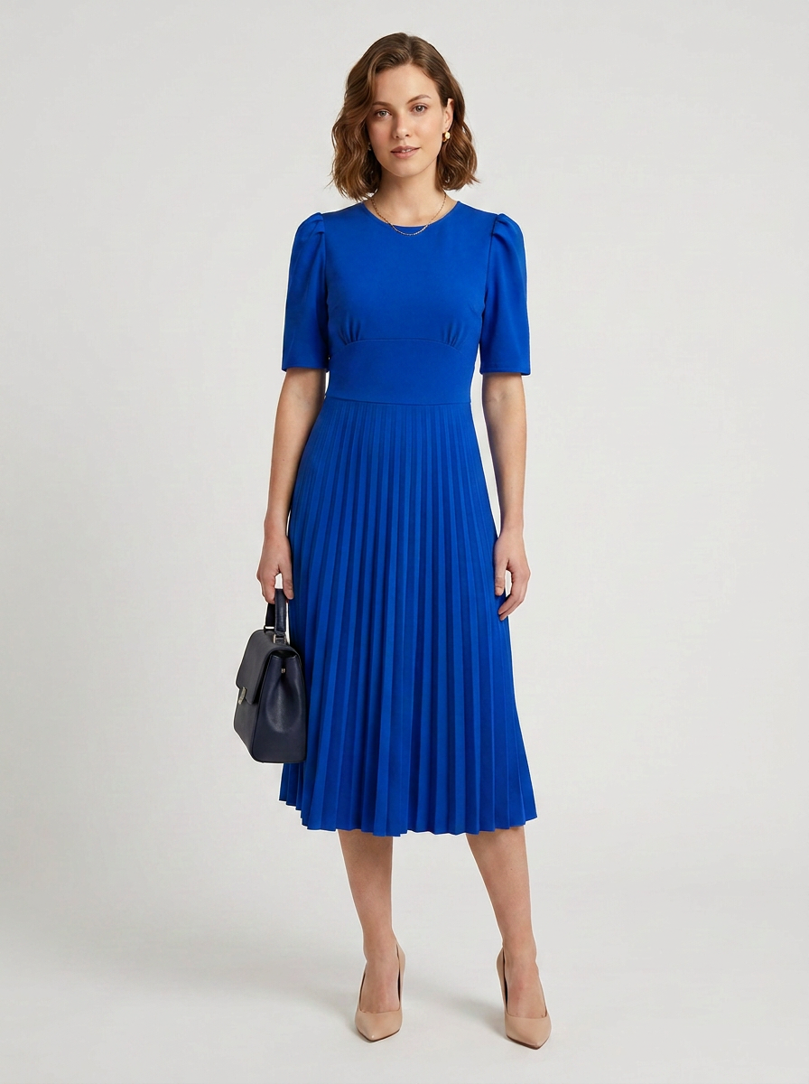 A woman modeling a bright royal blue midi dress with short puff sleeves and a pleated skirt, holding a small black handbag against a light gray studio background.
