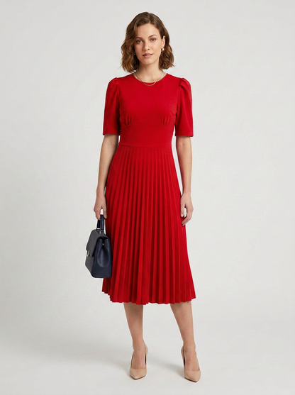 A woman modeling a bright red midi dress with short puff sleeves and a pleated skirt, holding a dark blue handbag against a light gray studio background.