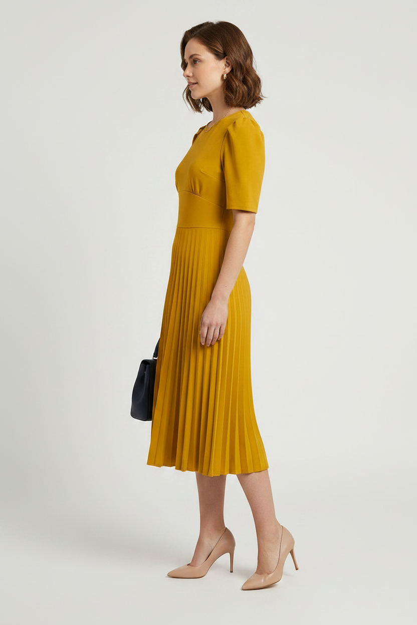 A woman modeling a mustard yellow midi dress with short puff sleeves and a pleated skirt, holding a small dark blue handbag against a light gray studio background.