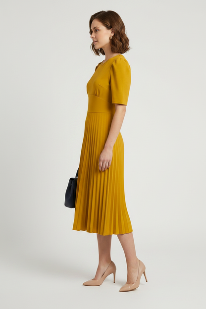 A woman modeling a mustard yellow midi dress with short puff sleeves and a pleated skirt, holding a small dark blue handbag against a light gray studio background.