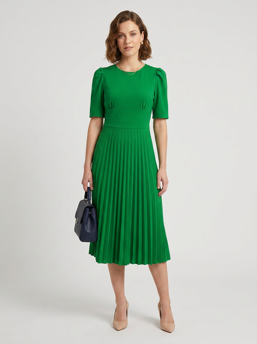A woman modeling a bright green midi dress with short puff sleeves and a pleated skirt, holding a dark navy handbag against a light gray studio background.