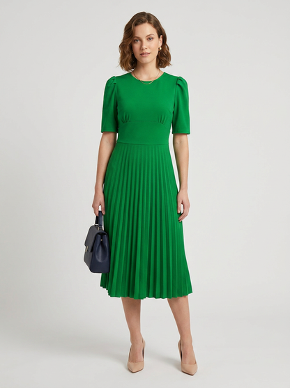 A woman modeling a bright green midi dress with short puff sleeves and a pleated skirt, holding a dark navy handbag against a light gray studio background.