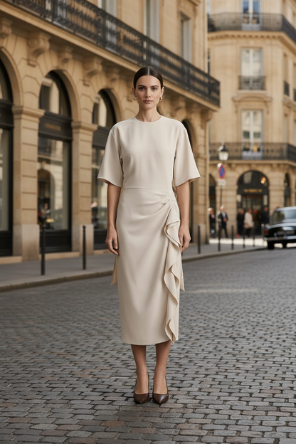 A woman stands on a cobblestone street wearing a light beige midi dress with short sleeves, a high neckline, and a draped asymmetrical ruffle detail at the skirt. The background shows classic Parisian-style buildings.