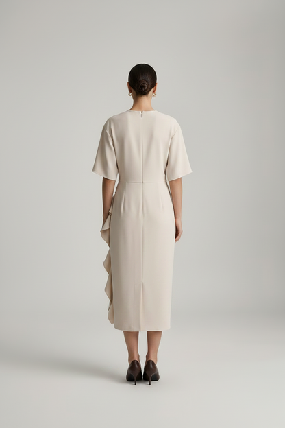 A model stands against a light gray studio background wearing a cream-colored midi dress with short sleeves, a round neckline, and a ruffled side detail, paired with dark brown pointed-toe heels.