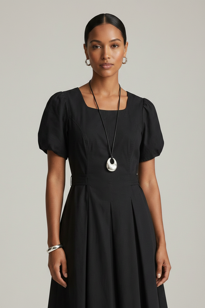 A woman modeling a black midi dress with short puff sleeves, a square neckline, and a flared skirt, photographed against a plain light gray studio background.