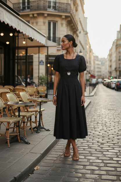 A woman stands on a cobblestone street wearing a black midi dress with puff sleeves, a square neckline, and a flared skirt. The background shows Parisian-style buildings and outdoor cafe seating.