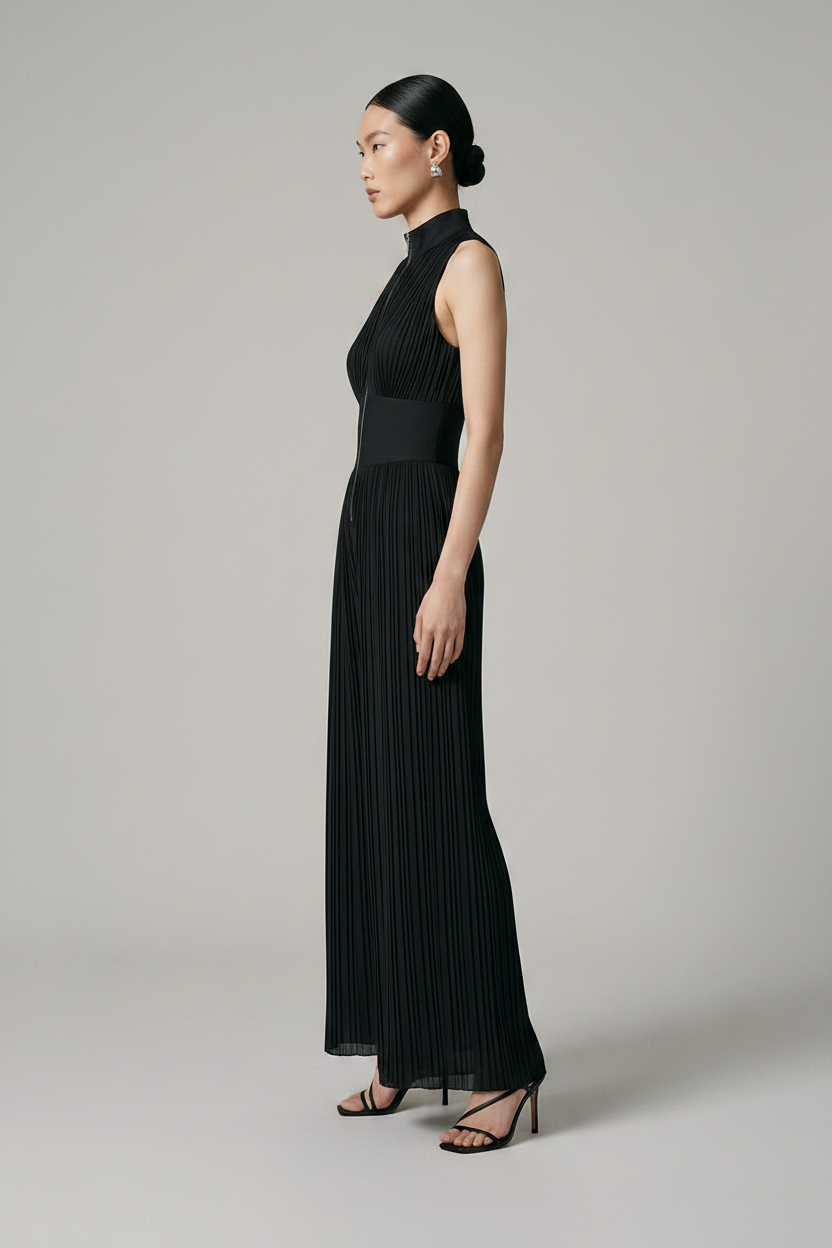 A model stands against a neutral light gray studio background wearing a sleeveless black pleated jumpsuit with a high mock neck, front zipper, and wide legs, paired with black strappy heels.
