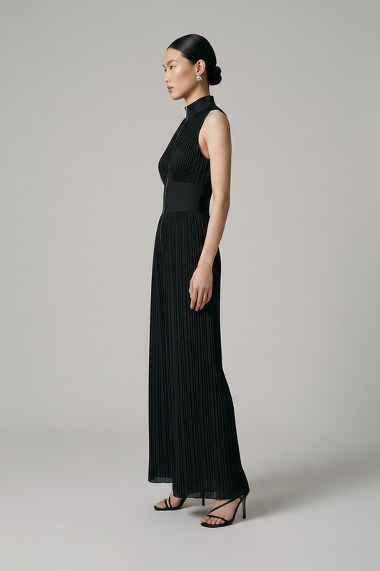 A model stands against a neutral light gray studio background wearing a sleeveless black pleated jumpsuit with a high mock neck, front zipper, and wide legs, paired with black strappy heels.