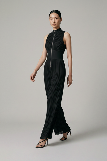 A model stands against a neutral light gray studio background wearing a sleeveless black pleated jumpsuit with a high mock neck, front zipper, and wide legs, paired with black strappy heels.
