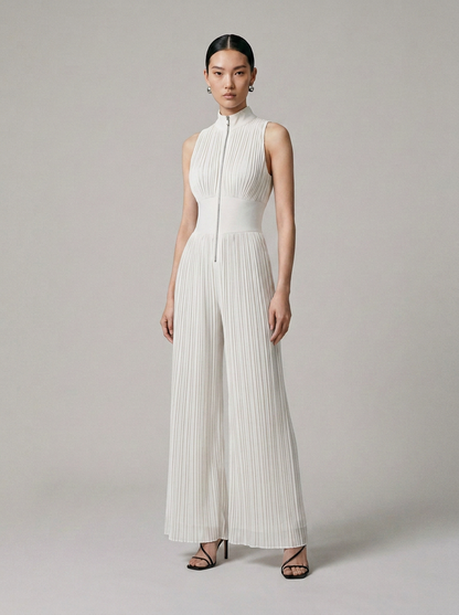 A model stands against a light gray studio background wearing a sleeveless white pleated jumpsuit with a high mock neck and front zipper, paired with black strappy heels.