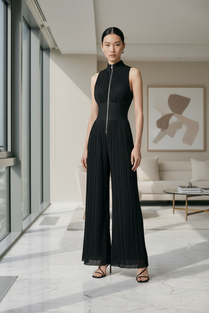 A model stands against a neutral light gray studio background wearing a sleeveless black pleated jumpsuit with a high mock neck, front zipper, and wide legs, paired with black strappy heels.