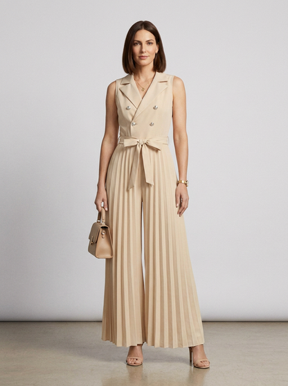 A woman models a sleeveless beige double-breasted jumpsuit with wide legs, pleated detailing, and a matching waist tie. She is holding a matching beige handbag and wearing beige high-heeled sandals against a neutral studio backdrop.