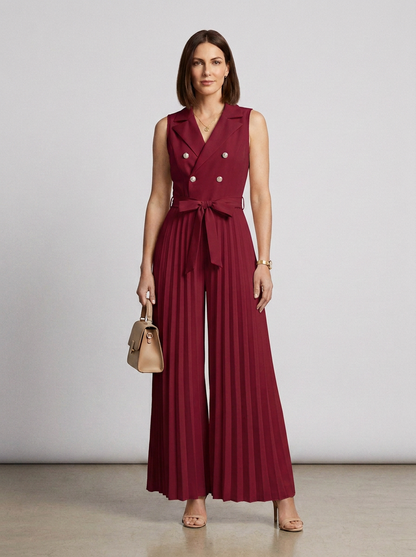 A woman models a sleeveless burgundy wide-leg jumpsuit with pleated detailing, double-breasted buttons, and a matching waist tie, holding a small beige handbag against a neutral studio background.