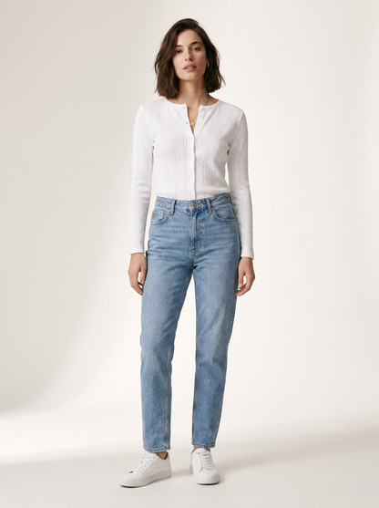 A woman modeling a white ribbed long-sleeve button-front top and light-wash blue jeans with white sneakers, standing against a plain off-white studio background.