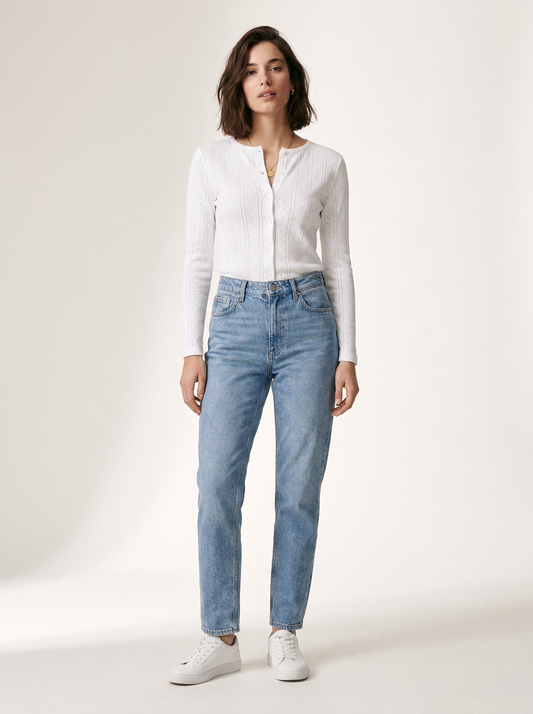A woman modeling a white ribbed long-sleeve button-front top and light-wash blue jeans with white sneakers, standing against a plain off-white studio background.