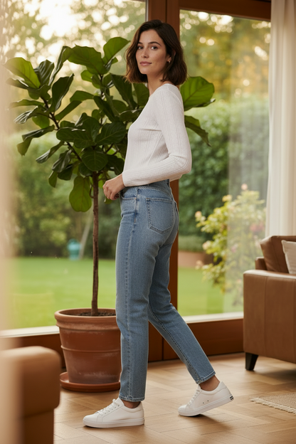 A woman stands in a bright living room wearing a white ribbed long-sleeve top, light blue high-waisted straight-leg jeans, and white sneakers, with a large potted fiddle-leaf fig and large windows in the background.