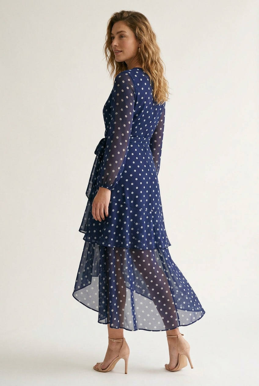 A woman modeling a navy blue long-sleeve midi dress with a white polka dot pattern, featuring a wrap-style V-neck bodice, tie waist, and a layered ruffled hem, set against a plain light background.