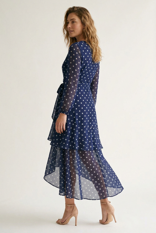 A woman modeling a navy blue long-sleeve midi dress with a white polka dot pattern, featuring a wrap-style V-neck bodice, tie waist, and a layered ruffled hem, set against a plain light background.