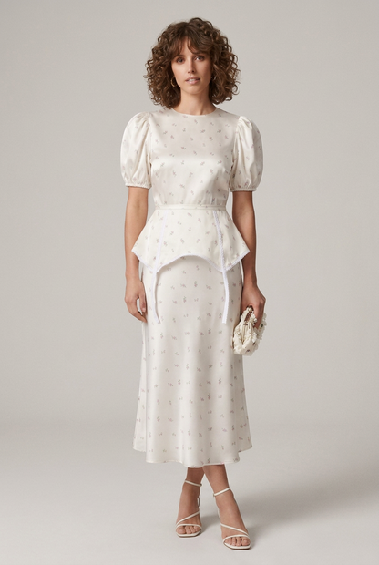 A woman models a white puff-sleeve midi dress with a delicate floral print, featuring a peplum waist and tie detail at the front. She is styled with white strappy heels and a matching white clutch, set against a plain light gray studio background.