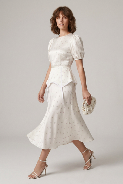 A woman models a white puff-sleeve midi dress with a delicate floral print, featuring a peplum waist and tie detail at the front. She is styled with white strappy heels and a matching white clutch, set against a plain light gray studio background.