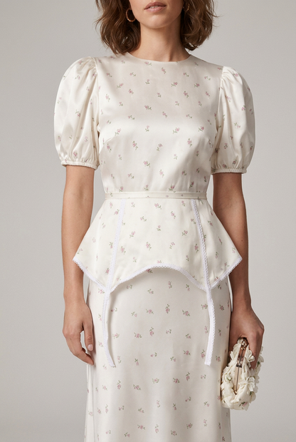 A woman models a white puff-sleeve midi dress with a delicate floral print, featuring a peplum waist and tie detail at the front. She is styled with white strappy heels and a matching white clutch, set against a plain light gray studio background.